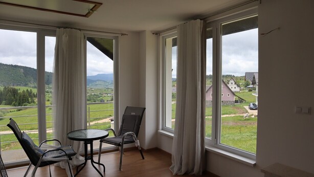 A balcony with a table and chairs, large windows with curtains, and a view of a green landscape and buildings. - Lotus Villas (Mudrike)