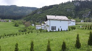 A white building with a sloped roof surrounded by a lush green field and a forested hill in the background. - Lotus Villas (Mudrike)