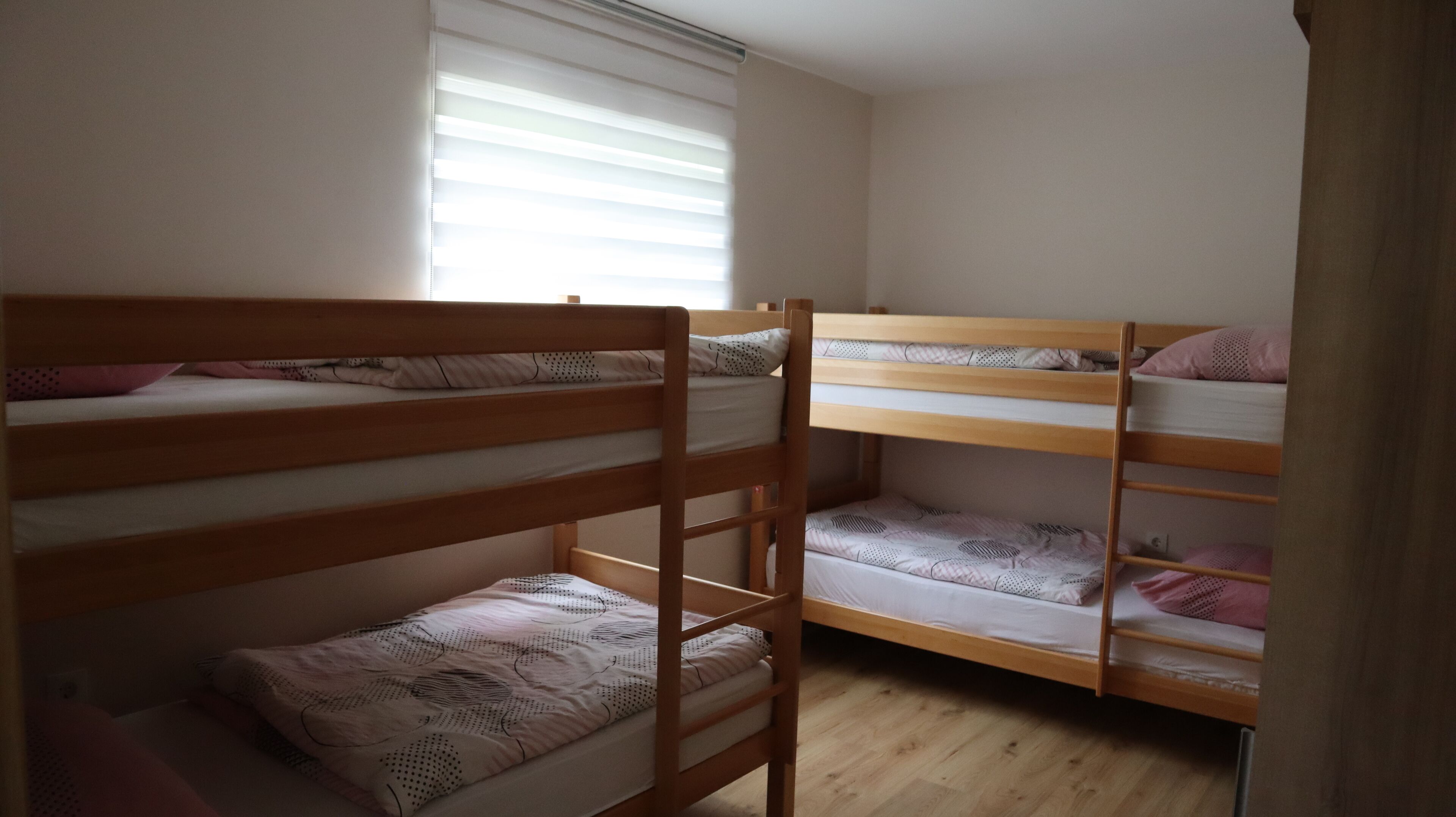 A dormitory room with bunk beds, wooden flooring, and a window with blinds.