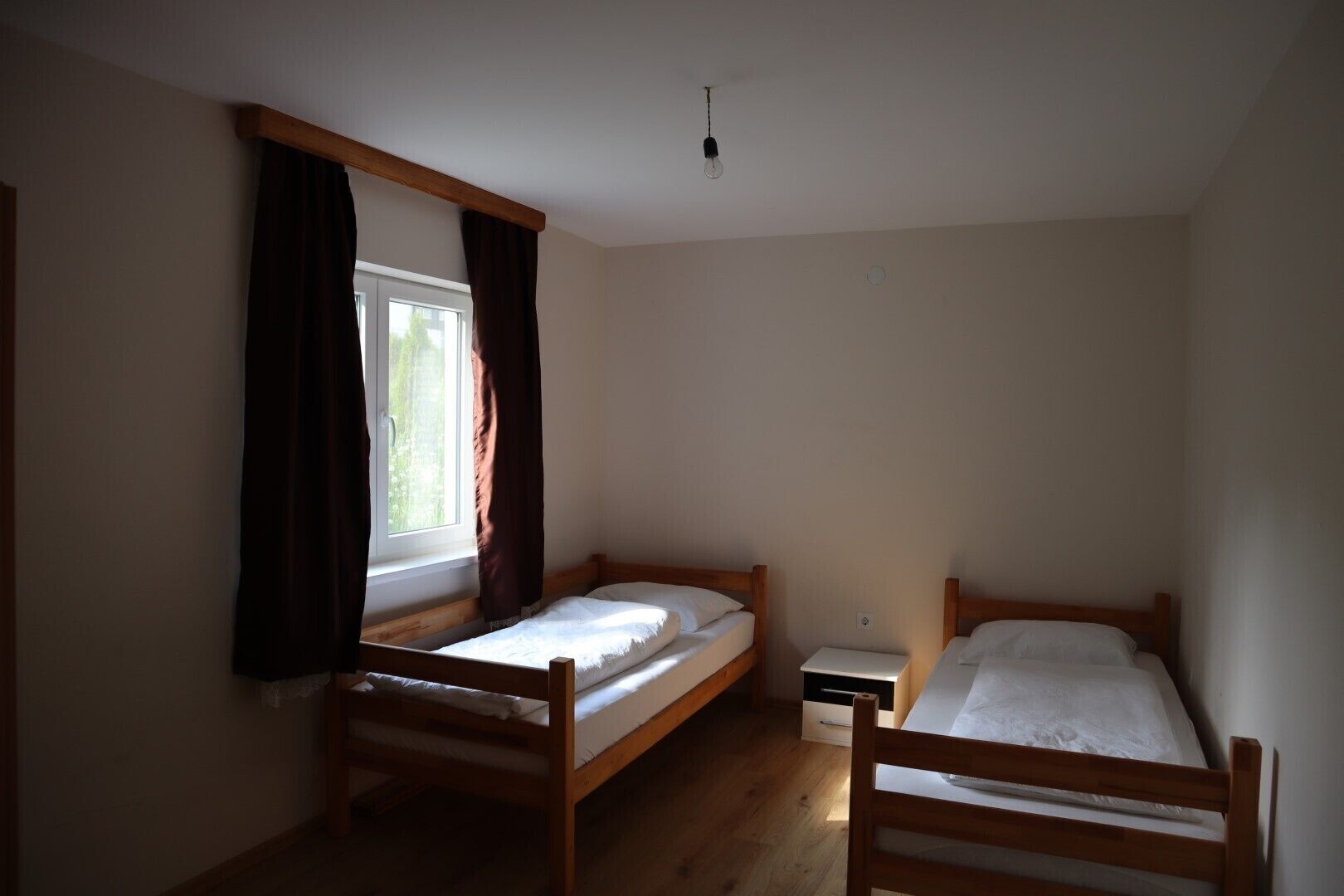 Een kamer met twee houten bedden, een klein nachtkastje, een raam met gordijnen en een lamp.