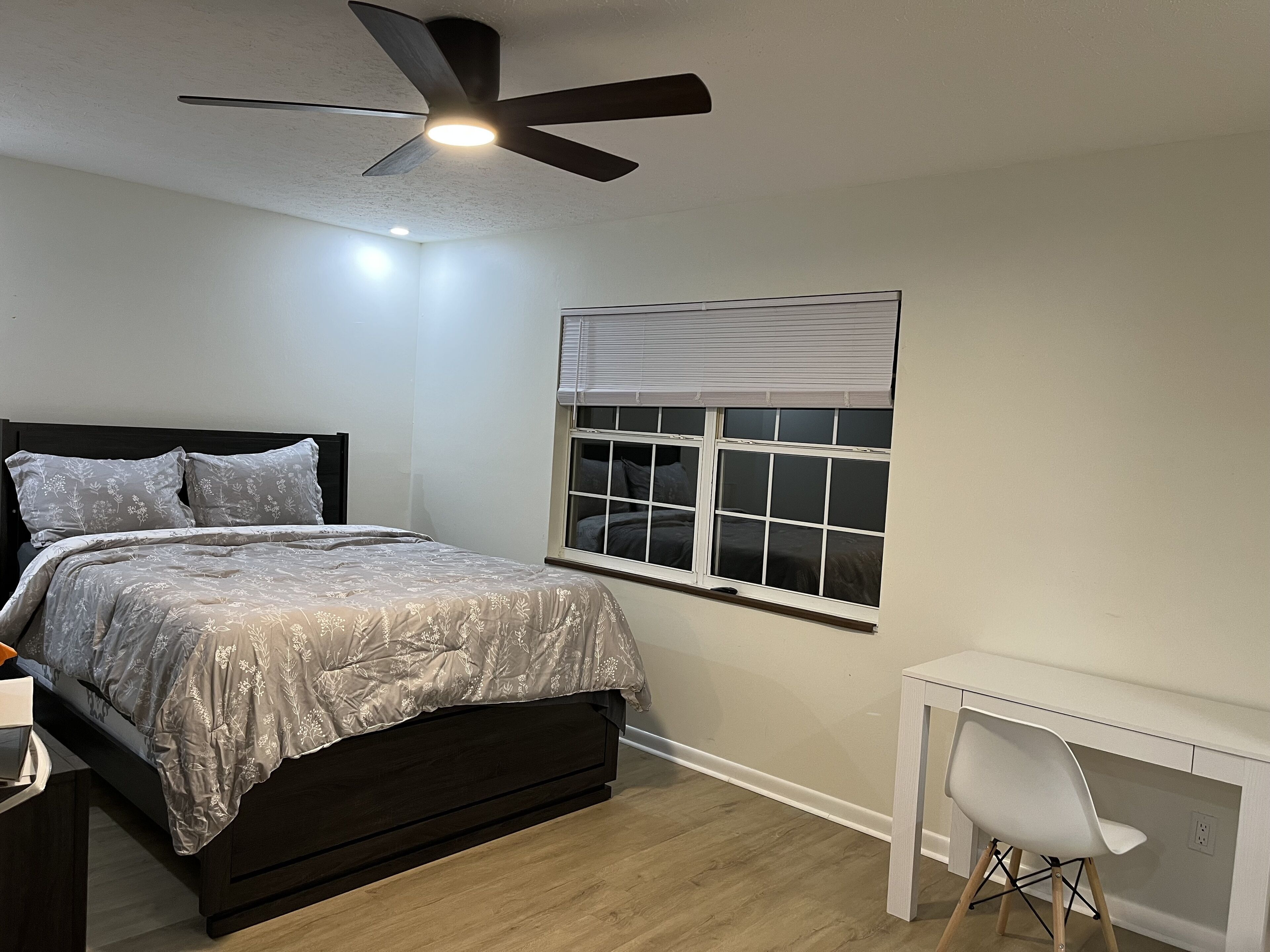A bedroom with a bed, a desk, a chair, a ceiling fan, and a window with blinds.
