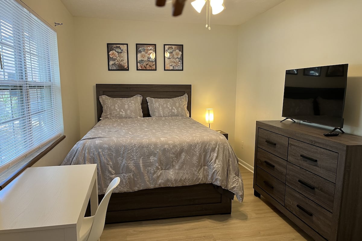 Ein Schlafzimmer mit einem Bett, einem Fernseher auf einer Kommode, einem Schreibtisch mit Stuhl und einem Fenster mit Jalousien.