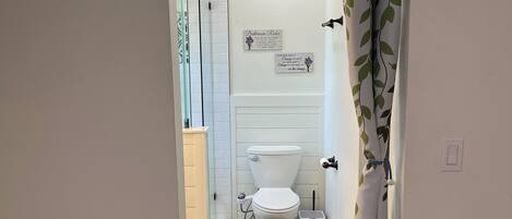 A bathroom with a toilet, bidet, and a small trash bin. There is a blue rug on the floor and a white wall with a light fixture.