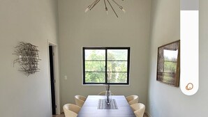A dining area with a dark wooden table, six chairs, a modern chandelier, a window with a view of greenery, and a wall-mounted abstract art piece.