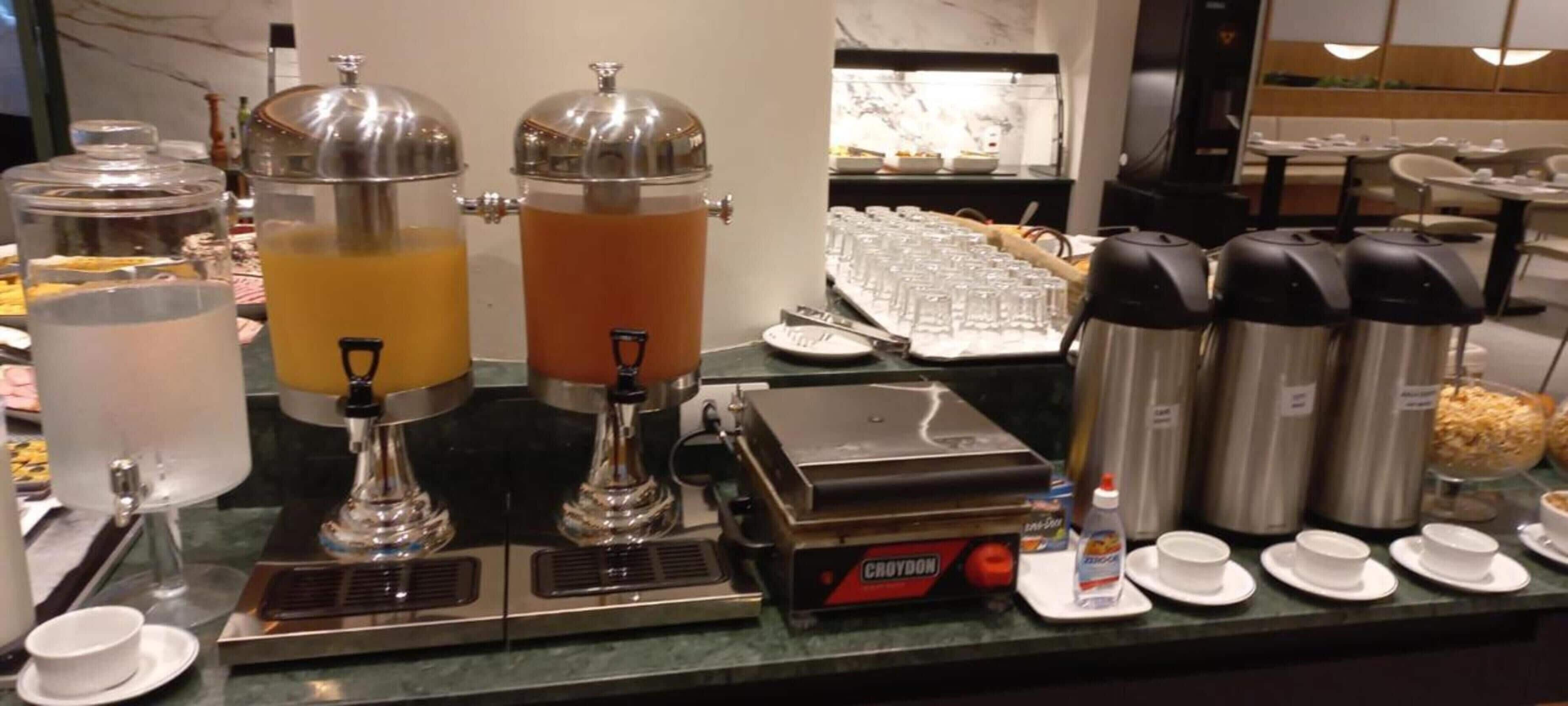 A buffet setup with various food and drink dispensers, including a juice cooler, a coffee station, and a buffet line with plates and glasses.