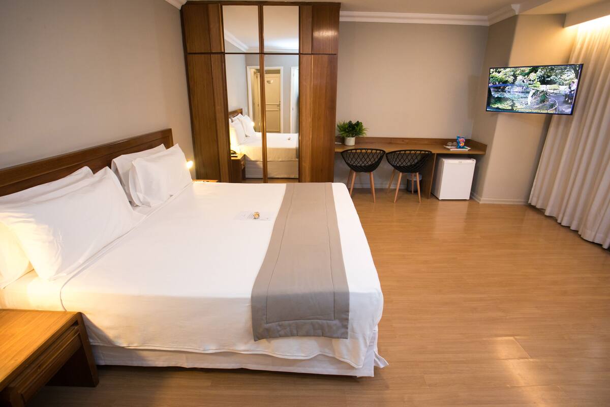 A hotel room with a large bed, a desk, two chairs, a small refrigerator, and a TV on the wall.