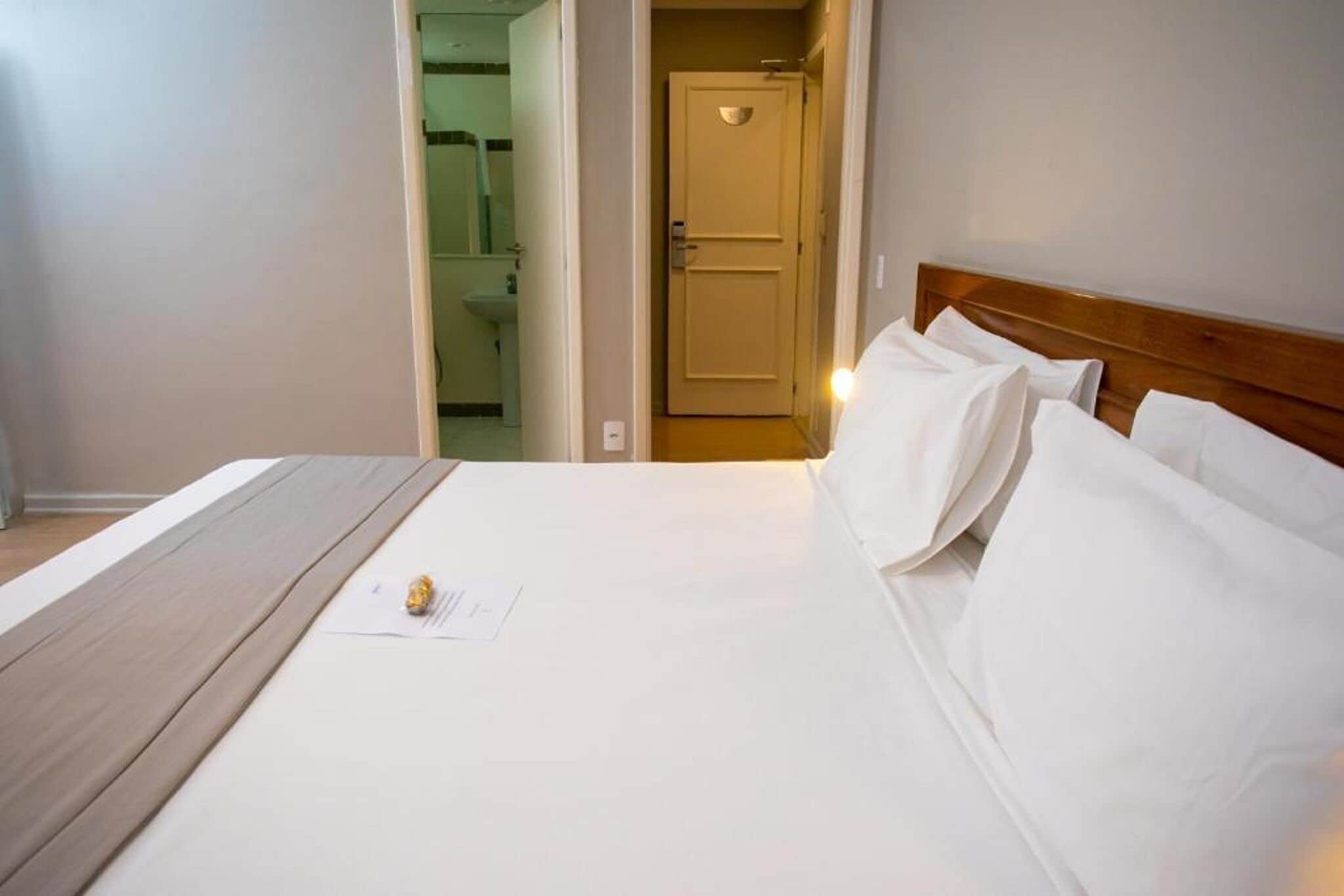 Habitación de hotel con cama, cabecera de madera, una puerta y un baño visible a través de un acceso abierto.