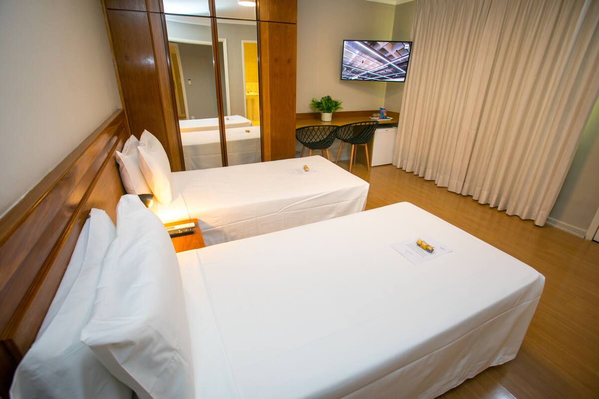 A hotel room with two single beds, a desk with a chair, a TV on the wall, and a large window with curtains.