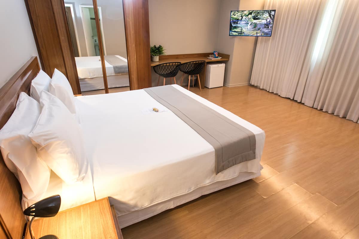 A neatly made bed with white linens and a gray throw blanket, a wooden nightstand with a lamp, a desk with two chairs, and a TV mounted on the wall.