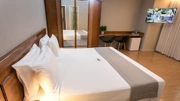 A neatly made bed with white linens and a grey throw, a small desk with a chair, and a television mounted on the wall.