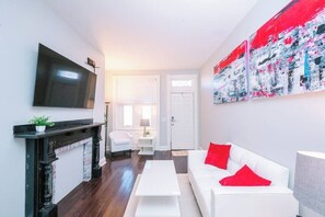 Una sala de estar con chimenea, un televisor de pantalla plana montado en la pared, un sofá blanco con cojines rojos y un cuadro abstracto de gran tamaño.