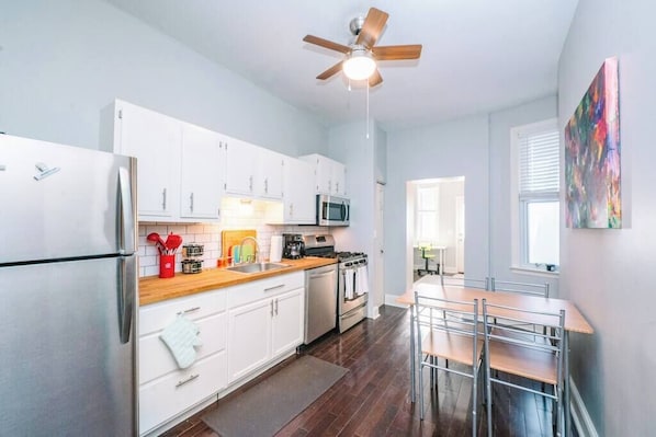A modern kitchen with white cabinets, a stainless steel refrigerator, a microwave, and a stove. There is a ceiling fan, a dining table with chairs, and a window with blinds.
