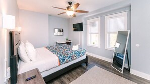 Un dormitorio con una cama, ventilador de techo, televisor, espejo y ventana.