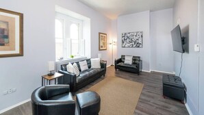 A modern living room with a sofa, armchair, ottoman, and a flat-screen TV mounted on the wall. - ROKU TVs - Washer & Dryer - QueenBeds - GroupStay (Pittsburgh)
