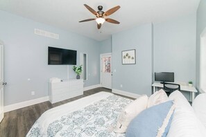 A bedroom with a bed, a ceiling fan, a TV, a desk, and a door.