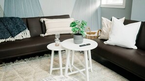 Ein Wohnzimmer mit einem braunen Sofa, zwei weißen Beistelltischen, einer Topfpflanze, einer Fernbedienung und einem Glas auf einem der Tische.