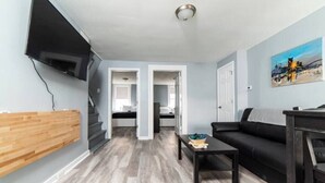 A modern living room with a black sofa, a wooden accent wall, a flat-screen TV mounted on the wall, and a staircase leading to another room. - -- AGH Walkable - Free Parking - Washer & Dryer -- (Pittsburgh)