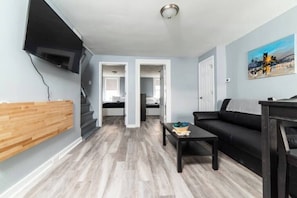 Un salon moderne avec un canapé noir, un mur d’accent en bois, un téléviseur à écran plat fixé au mur et un escalier menant à une autre pièce.