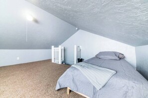 Um quarto com teto inclinado, uma cama com colcha cinza e piso coberto de carpete.
