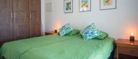 A bedroom with a large wooden wardrobe, a green bed with leaf-patterned pillows, and three framed artworks on the wall.