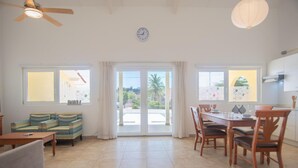 A spacious living area with a sofa, dining table, and chairs. There is a ceiling fan, a clock, and large windows with curtains. - NEW Casa Orcuyo in Santa Cruz (Santa Cruz)