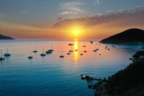 A coastal sunset with boats on calm waters, rocky coastline, and a distant hill.