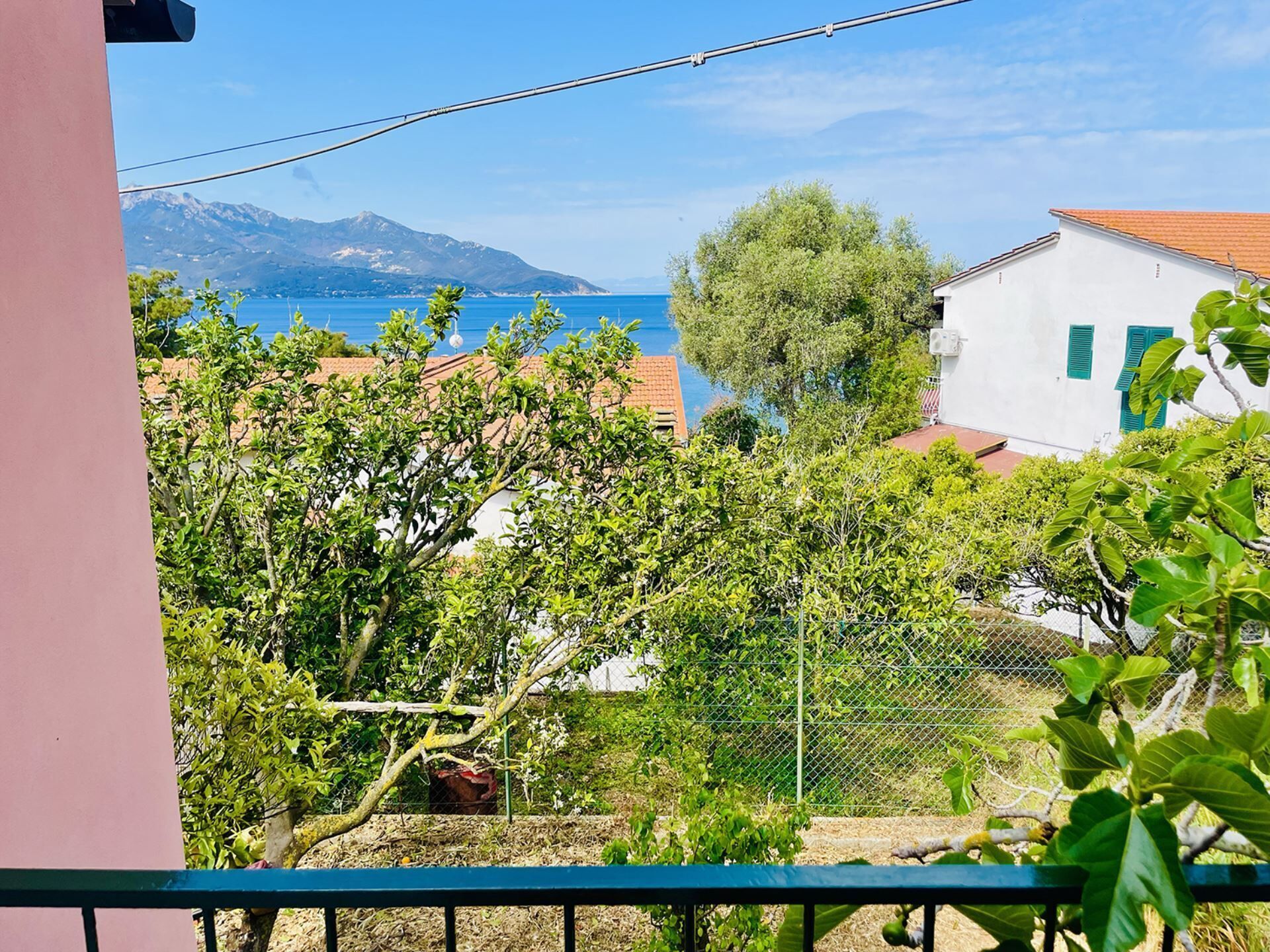 A balcony with a view of a coastal town, a mountain in the distance, and a fence with greenery.
