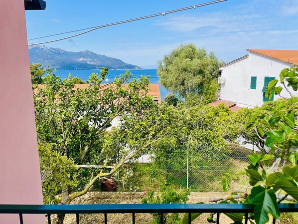 A balcony with a view of a coastal town, a mountain in the distance, and a fence with greenery.