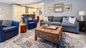 Una sala de estar con un sofá gris, un sillón azul y un reposapiés marrón. Se ve una cocina al fondo.