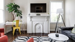 A living room with a fireplace, a TV mounted above it, a desk with a chair, a large plant, and a sofa. - Modern, Convenient Gem: Urban Retreat 'n Richmond (Richmond)