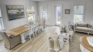 A spacious living room with a wooden dining table, white chairs, a beige sofa, and a small side table.