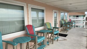 A covered outdoor seating area with multiple Adirondack chairs and small tables, a building with large windows, and a visible door. - Beachgate Condo Suites And Hotel 231 1 Bedroom Condo (Port Aransas)