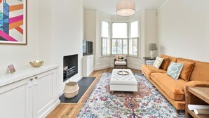 A living room with a sofa, ottoman, and a fireplace. There is a television, a lamp, and a framed picture on the wall. - Family Home near Clapham Common (London)
