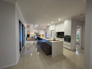 A modern kitchen with a central island, stainless steel appliances, and a dining area with a view. - Lily Pad Beach House (Agnes Water)
