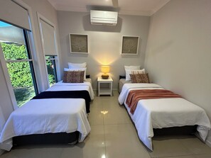 A hotel room with two beds, a small table with a lamp, and a window with a view of greenery. - Lily Pad Beach House (Agnes Water)