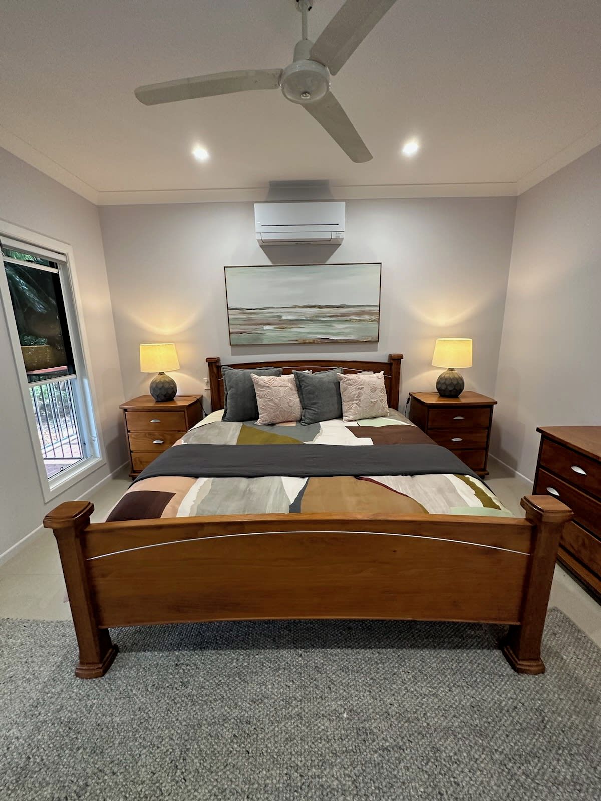 A bedroom with a bed, two bedside tables with lamps, a ceiling fan, an air conditioning unit, and a window with a view.