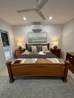 A bedroom with a bed, two bedside tables with lamps, a ceiling fan, an air conditioning unit, and a window with a view. - Lily Pad Beach House (Agnes Water)