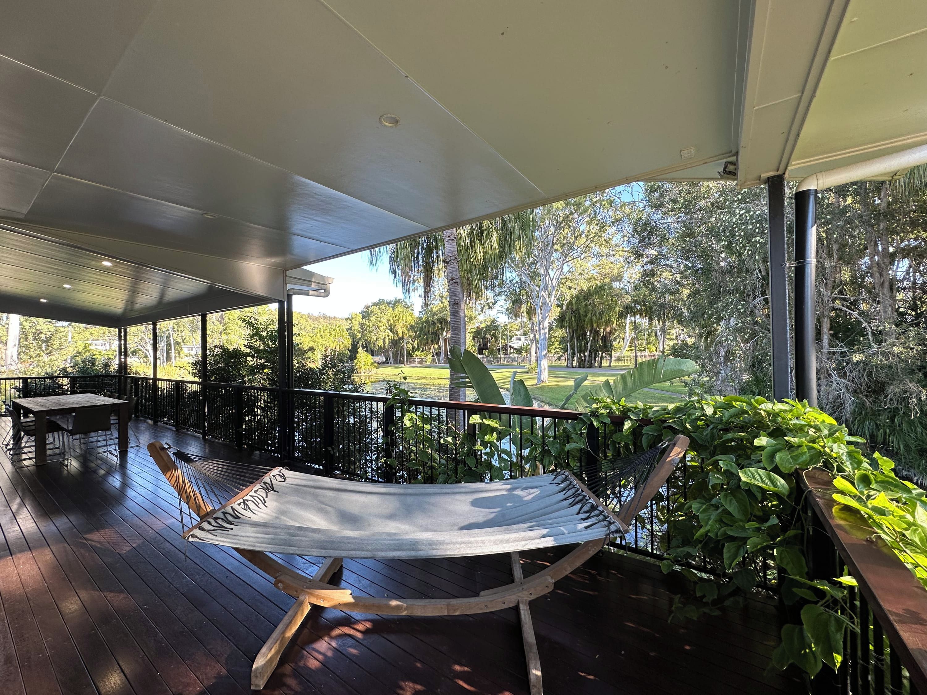 A covered outdoor area with a hammock, wooden deck, and a view of a park with trees and a body of water.