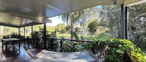 A covered outdoor area with a hammock, wooden deck, and a view of a park with trees and a body of water.