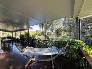 A covered outdoor area with a hammock, wooden deck, and a view of a park with trees and a body of water. - Lily Pad Beach House (Agnes Water)