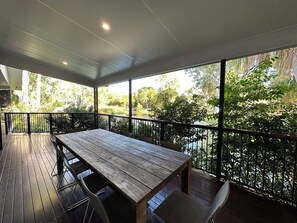 A covered outdoor area with a wooden table and chairs, surrounded by a metal railing, overlooking a lush garden. - Lily Pad Beach House (Agnes Water)