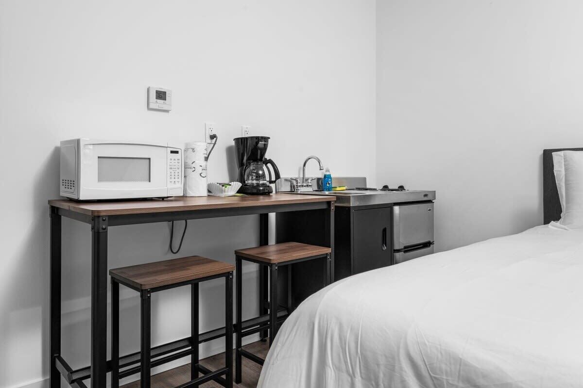 A compact hotel room with a bed, a kitchenette with a microwave, coffee maker, and a small refrigerator. There are two stools at the kitchenette.