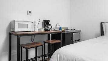 A compact hotel room with a bed, a kitchenette with a microwave, coffee maker, and a small refrigerator. There are two stools at the kitchenette.