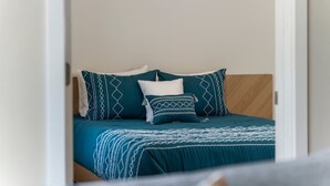 Una cama con colcha color turquesa y almohadas blancas, cabecera de madera y pared lisa.