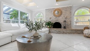 Un salón amplio con un sofá blanco, una mesa redonda con una planta, una chimenea de ladrillo y ventanas grandes.