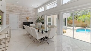 Un comedor amplio con una mesa de vidrio y sillas blancas, una cocina con encimera negra y vistas a un jardín trasero con piscina.