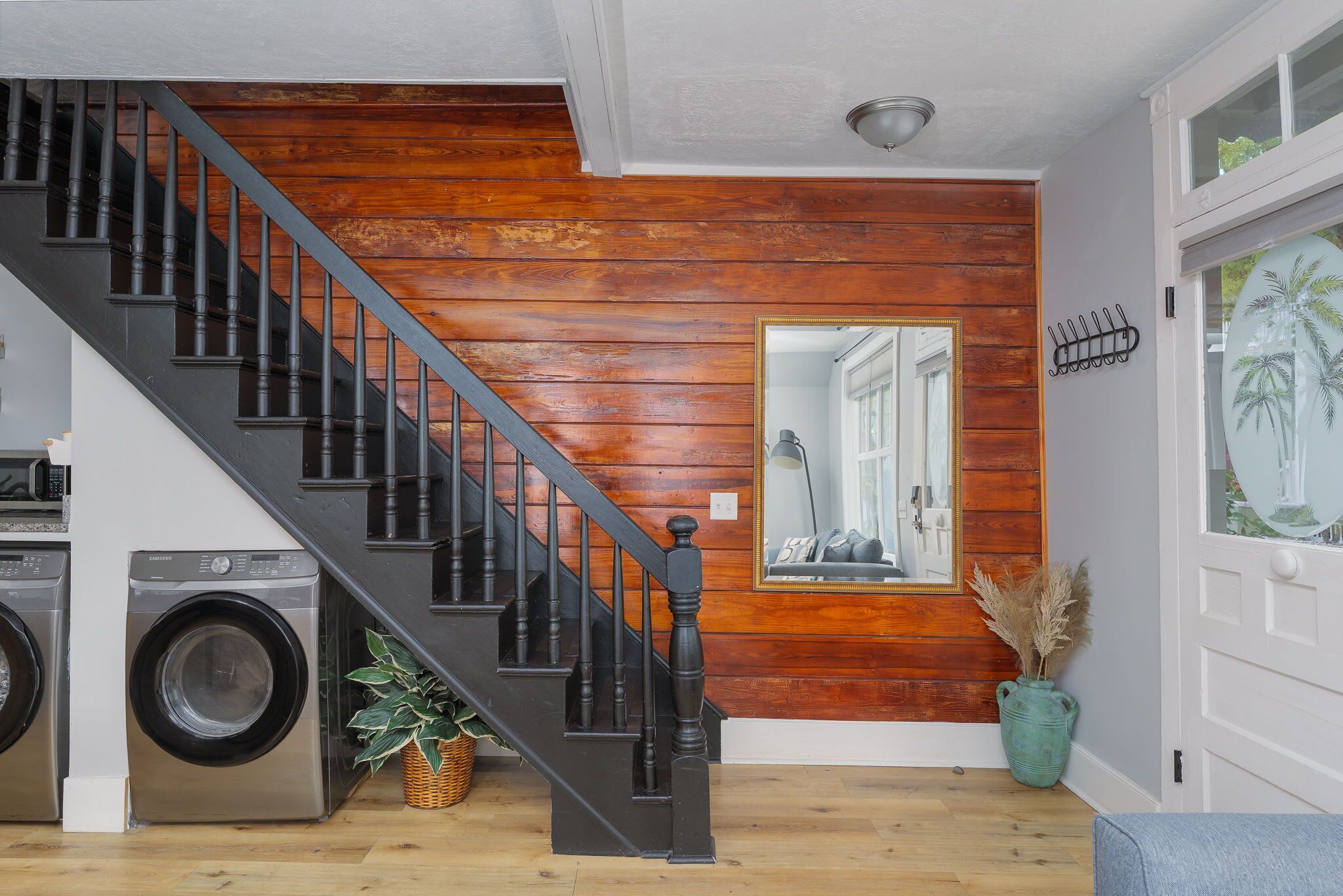 A staircase with black balusters leading to a wooden wall, a washing machine, a potted plant, and a door with a decorative mirror.