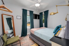 Een slaapkamer met een bed, een stoel, een bureau, een spiegel, een plafondventilator en een raam met gordijnen.