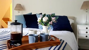Ein ordentlich bezogenes Bett mit einer blau-weiß gestreiften Tagesdecke, ein Nachttisch mit Lampe, ein Tablett mit Kaffeekanne, Tassen und einer Blumenvase.