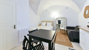 Une pièce compacte et bien éclairée, comprenant un lit, une petite table à manger avec des chaises, un canapé et un coin cuisine.